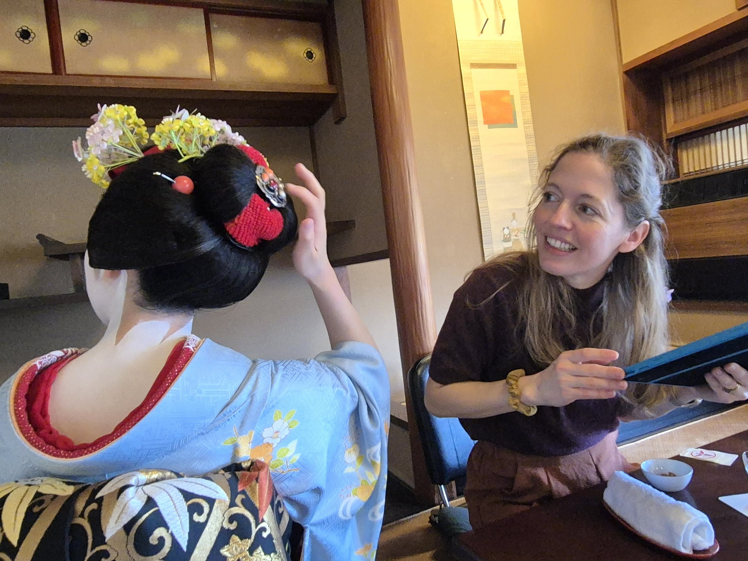 A smiling tourist sits across from a geisha in a blue kimono and ornate hairstyle inside a traditional Japanese room, observing and interacting closely during a cultural experience.