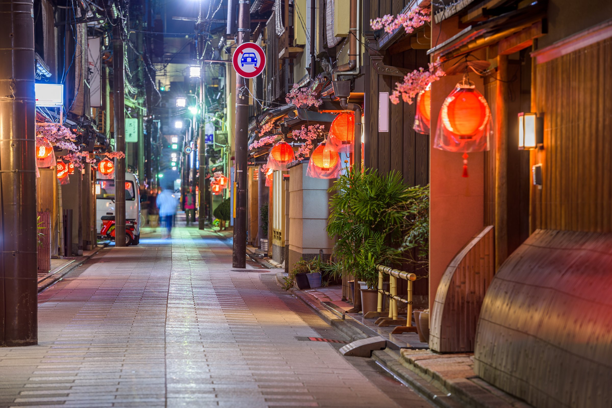 A quiet, lantern-lit alley in Kyoto at night, lined with traditional wooden buildings adorned with red lanterns and artificial cherry blossoms, with a blurred figure walking in the distance.
