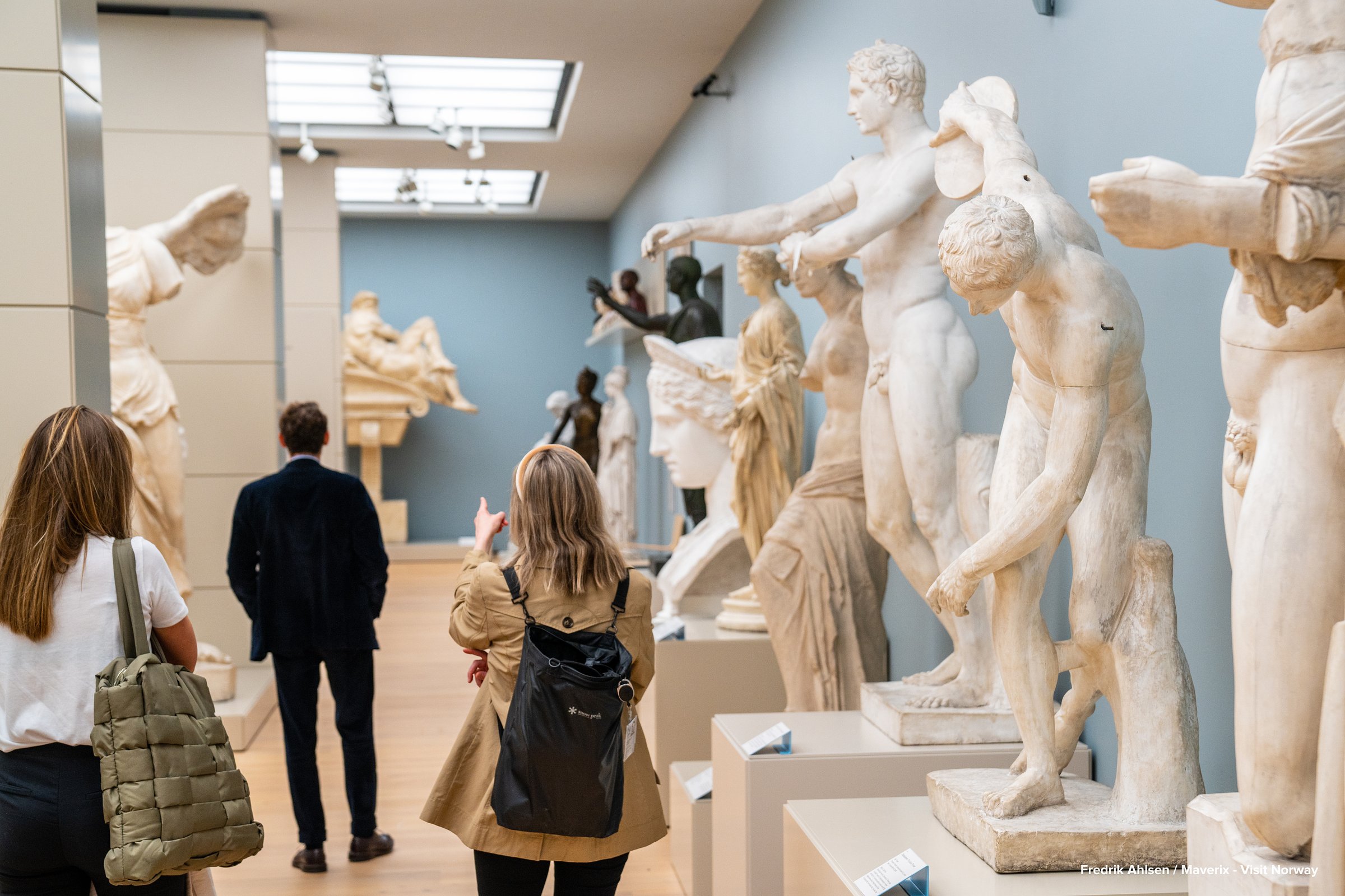 Visitors walk among classical marble statues in a bright exhibition hall.