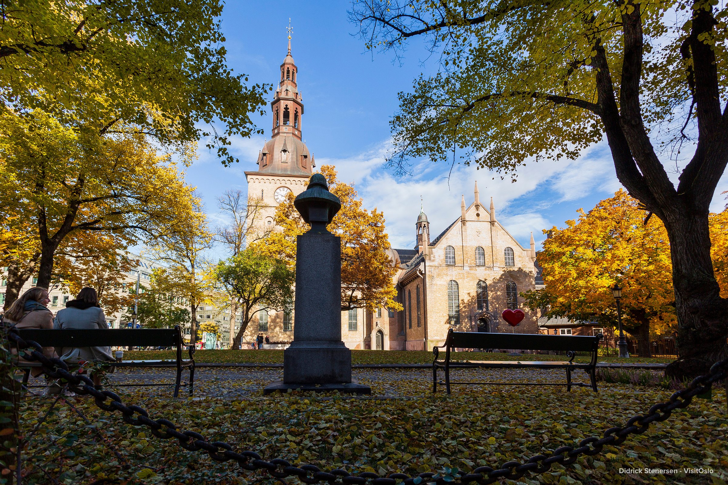 Brick church framed by golden trees and park benches under a clear blue sky.