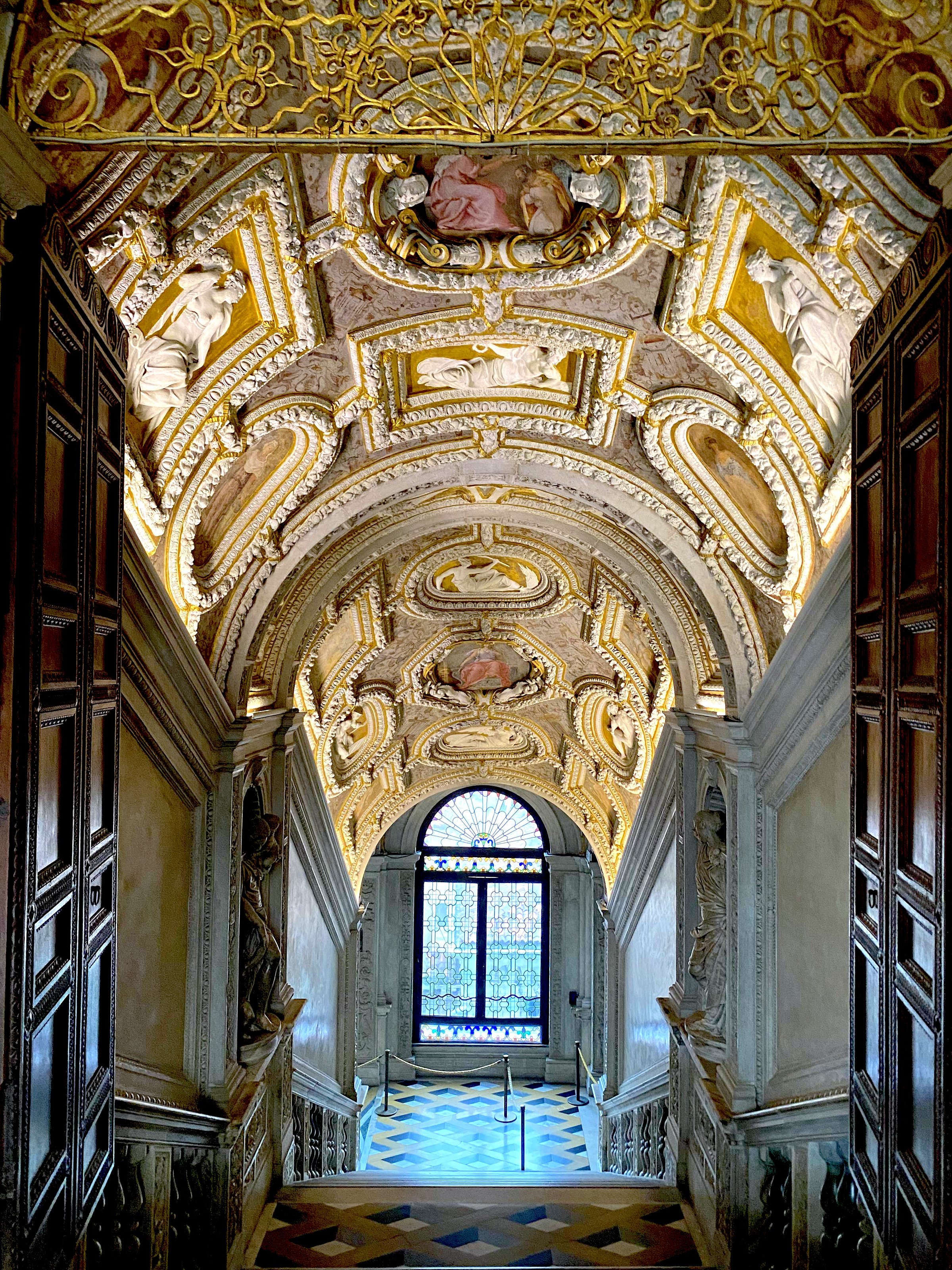 Ornate staircase inside the Doge's Palace in Venice, featuring a richly decorated vaulted ceiling with gold stucco, sculptures, and a stained-glass window at the bottom.