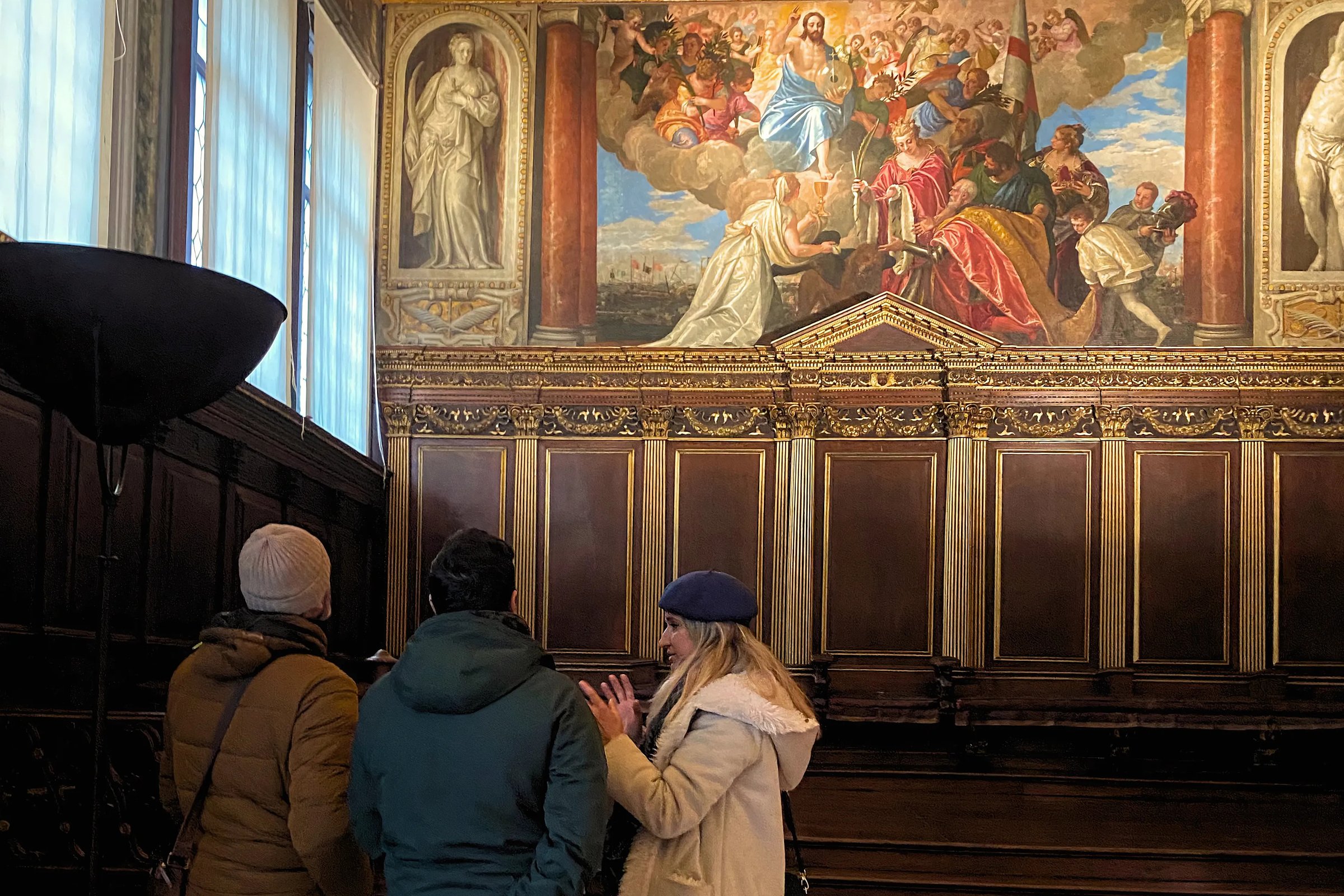 A small group listens to a guide inside the Doge’s Palace, standing before a large Renaissance painting depicting a religious and ceremonial scene surrounded by ornate wood paneling.